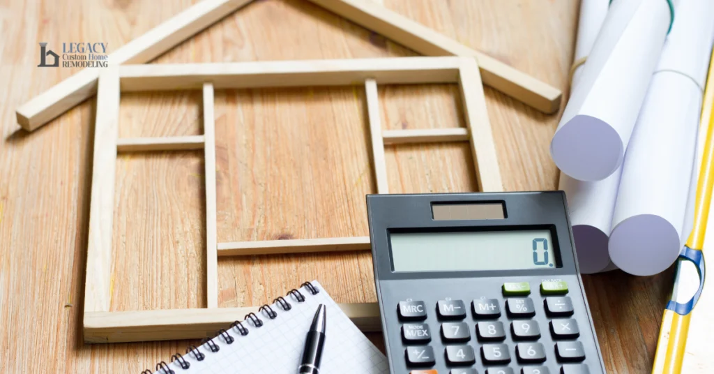 Wooden table with house frame model, calculator showing zero, rolled architectural plans, notebook, and pen, conveying planning and construction theme.