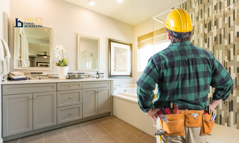 A construction worker in a yellow helmet and tool belt stands in a modern bathroom. He faces a bathtub, gray cabinets, and decorative tiles, conveying a sense of renovation and craftsmanship.
